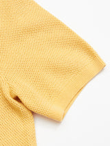 A close-up of the Filo Knit Polo in Yellow Grain by Blue de Gênes shows its yellow cotton sweater sleeve with a textured waffle pattern and ribbed cuff, set against a plain white background. The fabric looks soft and slightly stretchy.