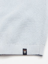Close-up of the Filo Knit Polo - Blue Water by Blue de Gênes, a light blue sweater made from 100% cotton with a retro repeat stitch pattern. Attached to the hem is a small black tag featuring orange and white logos.