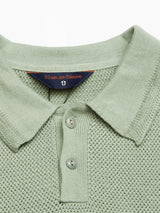 Close-up of the Filo Knit Polo - Green Bay by Blue de Gênes, a light green knitted polo shirt made from 100% cotton. It showcases a retro repeat stitch pattern, folded collar, short button placket with two buttons, and inside is a small navy label with Blue de Gênes in orange.