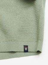 Close-up of the Filo Knit Polo in Green Bay by Blue de Gênes. This light green, 100% cotton sweater features a retro repeat stitch for timeless appeal, a textured pattern, and ribbed hem. A small black tag with crown and shield emblem is attached near the bottom edge.