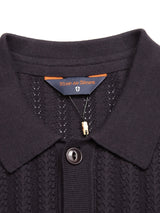 The Compra Open Polo Knit in Dark Navy by Blue de Gênes showcases a textured design with a collar, corozo nut buttons, and a tag at the neckline, from which hangs a small gold clip.