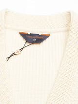 Close-up of the Cappa Stripe Cardigan in whitecap, a cream-colored knit made from soft cotton with a textured pattern. The inside label reads Blue de Gênes with a small shield emblem. A black string with a wooden tag adds an artisan touch.