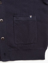 The Achille Cardigan - Navy by Blue de Gênes features intricate knitting techniques, a textured pocket, and ribbed hem. Corozo buttons line the left side, while a small emblem patch above the pocket adds a subtle flair.