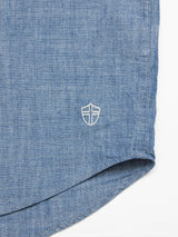 Close-up of the Sandro Chambray Shirt by Blue de Gênes, featuring light blue chambray fabric and a small white shield emblem on the bottom left. The button-down collar adds classic style, with textured stitching on the hem. Background is plain white.