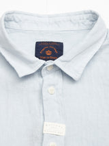 The Enrico Beach Shirt - Blue Water by Blue de Gênes is a light blue button-up summer shirt with a collar, showcased on a white background. Made from linen, it features Corozo nut buttons and has a visible black label near the collar with another below the buttons.