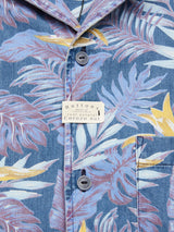 A close-up of the Blue de Gênes Resort Melfi Shirt—Multi Pattern reveals a blue tropical-patterned design with large leaves in white, pink, and yellow. Made from denim cotton, it features grey Corozo nut buttons and a small tag near the top button.