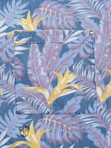 Close-up of the Resort Melfi Shirt by Blue de Gênes in a multi-patterned blue fabric featuring tropical leaves and subtle flowers in purple, yellow, and white. A pocket adorns the center, with a small embroidered logo on the bottom left, capturing its laid-back elegance.