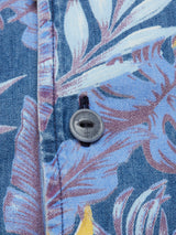 A close-up of Blue de Gênes Resort Melfi Shirt - Multi Pattern shows a blue floral fabric with large tropical leaves in lighter shades, accented by yellow. A dark button and stitching along the edge suggest its perfect for a casual, stylish look.