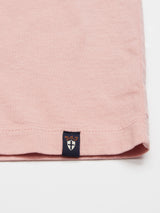 Close-up of the Salty Logo T-Shirt - Porcelain Pink by Blue de Gênes, displaying a pink cotton-jersey fabric corner with a small black tag featuring a white cross and gold text. Visible stitching along the hem on a plain white background, resembling fine chest embroidery on a crewneck.
