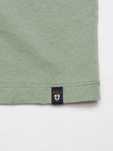 Close-up of the hem of the Salty Logo T-Shirt - Green Bay by Blue de Gênes, a light green cotton-jersey shirt. Features a small black tag with B.D.G in orange above a white shield emblem, complementing its regular fit.