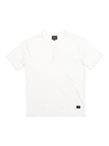 The BdG S/S Grandad in Snow White by Blue de Gênes is a cotton slub yarn, short-sleeve t-shirt with a buttoned placket using corozo nut buttons, accented by a black tag at the lower left corner against a plain white background.