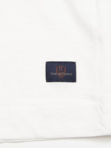 Close-up of the BdG S/S Grandad - Snow White t-shirt by Blue de Gênes, featuring a black square label with an orange True Gents emblem in cursive. Made from soft, lightweight cotton slub yarn, it merges comfort and style effortlessly.