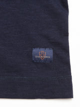 Close-up of Blue de Gênes BdG S/S Grandad - Navy fabric, featuring a dark blue cotton slub yarn with a small stitched label. The label displays a shield design and the text blue genes in orange and gray. The horizontal weave pattern resembles a classic grandad t-shirt style.