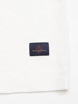 Close-up of a white fabric featuring a small, square black label with an orange emblem and the text Blues & Bones in white, showcasing intricate stitching and cotton slub yarn texture reminiscent of a classic grandad t-shirt from Blue de Gêness BdG Grandad - Snow White collection.