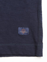 Close-up of navy cotton slub fabric with visible stitching, similar to a grandad t-shirt. A small rectangular tag features an orange emblem and text reading Blue de Gênes, crafted for a textured appearance. Product: BdG Grandad - Navy.