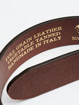 Close-up of the FlexCord Belt - Cognac by Blue de Gênes, made from brown full-grain leather with a Magikflex buckle and embossed gold text: Vegetable Tanned, Handmade in Italy. The light background highlights its texture and craftsmanship.