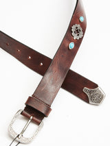 The Western Studded Belt by Blue de Gênes features dark brown full-grain leather with turquoise studs, silver embellishments, and an engraved silver buckle and tip.