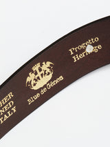 The Western Studded Belt in dark brown by Blue de Gênes features a close-up of embossed gold text: Leather Tanned Italy, Blue de Gênes, and Progetto Heritage, with a crest logo of a crown, castle, and two griffins. The design is elegantly accented with turquoise studs.