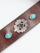Close-up of the Western Studded Belt - Dark Brown by Blue de Gênes. This hand-made Italian leather belt features an intricate silver centerpiece with swirling patterns flanked by two oval turquoise stones, elegantly displayed on a plain white surface.