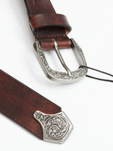 Close-up of the Western Studded Belt - Dark Brown by Blue de Gênes, featuring an ornate silver buckle and tip with intricate floral engravings for a vintage Western look. Hand-made in Italy, this full-grain leather belt showcases its texture and shine with partial curling.