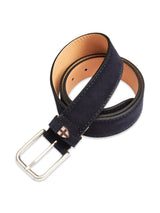The Suede Belt - Navy by Blue de Gênes, handmade in Italy, features a silver buckle, brown underside, and a cross-embroidered patch near the buckle. It is elegantly rolled up on a plain white background.