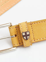 Close-up of the Suede Belt - Yellow Grain by Blue de Gênes, showcasing a metal buckle on the left. A shield emblem with a cross is attached to the belt loop. The background is light beige, partially showing another leather item.