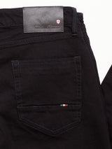 Close-up of the back pocket on Blue de Gênes Vinci Venom Blk Jeans - Dark Denim, with a black leather brand patch, dark stitching, a striped tag, and small red and white logo above. Made from black comfort stretch twill.