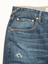 Close-up of Blue de Gênes Giacomo Osaka Jeans in dark denim, featuring yellow stitching, a small front pocket, a distressed thigh rip, and a tiny shield-shaped emblem near the pocket.