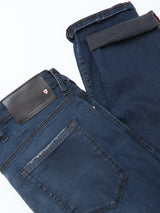 The Repi BlueBlk Used Jeans by Blue de Gênes feature a dark blue-black slim fit, laid flat with one leg folded. Details include a back pocket, belt loops, and a black leather patch with a red and white shield logo. Made from comfort stretch denim.