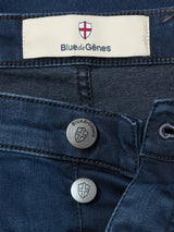 Close-up of Blue de Gênes Repi BlueBlk Used Jeans in blue-black stretch denim, with two shield-embossed metal buttons and a label above the waistband featuring a red cross and the Blue de Gênes brand name.