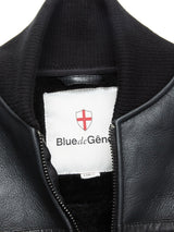 Close-up of the Rufo Shearling Jacket - Scarab by Blue de Gênes, featuring a black leather finish, ribbed collar, and partially unzipped to show a white Blue de Gênes label with a red shield emblem inside.