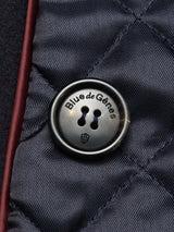 Close-up of a glossy button featuring the Blue de Gênes shield emblem, sewn onto dark navy Italian Melton wool with visible stitching and burgundy trim, highlighting the classic style of the Shiba Melton Coat by Blue de Gênes.