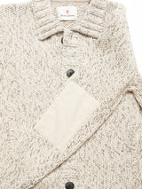 A close-up of the Sakura Knit Cardigan - Stone by Blue de Gênes, a beige speckled wool blend with dark buttons, chunky knit texture, collar, sleeve patch, and the Blue de Gênes label visible at the top.