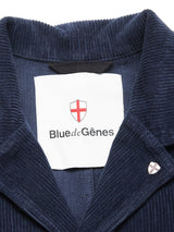 Close-up of the Masami Cord Blazer - Navy by Blue de Gênes. This garment-dyed corduroy blazer features a white brand label with a red cross emblem and a matching emblem pin on the lapel.