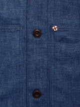 Close-up of the Blue de Gênes Yuma Fano Jacket in Denim Blue, a heavy cotton blend with two black buttons, two pockets, and a shield-shaped enamel pin featuring a red cross on white.