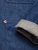 Close-up of the Yuma Fano Jacket in Denim Blue by Blue de Gênes, featuring workwear style, front pocket, a red cross pin on a white shield, and turned-up sleeve cuffs revealing striped inner fabric.
