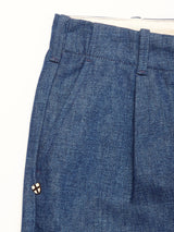 Close-up of Blue de GĂȘnes Shogun Fano Pant in denim blue with pleats, waistband belt loops, and a small round pinâwhite with a black cross and red detailânear the pocket, crafted from heavy cotton blend.