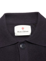 Close-up of a navy textured collared shirt with a black button and white Blue de Gênes label, perfect to pair with the Gifu Knit Cardigan - Navy from Blue de Gênes. A red shield logo sits above the text.
