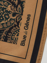 A close-up of the Siruku Scarf - Mixed Colours from Blue de Gênes shows an intricate black and teal floral pattern on tan, with the brand’s name printed in black along the edge of this stylish flower print scarf.