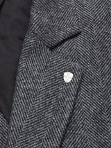 Close-up of the Fukui Blazer - Black Pattern by Blue de Gênes, a gray wool herringbone jacket with relaxed tailoring and a cross-design shield lapel pin on the left; its black inner lining is partially visible.