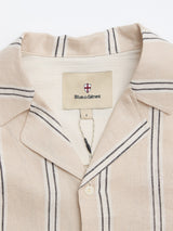 A close-up of the BGcody Fling Shirt in Pale Khaki by Blue de Gênes, featuring black and white vertical stripes. The oversized linen shirt is buttoned at the top, with a collar label displaying the brand name and shield logo.