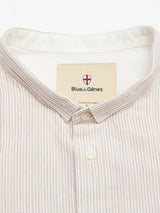 A close-up of the BGbasso Meco Shirt - Beige Stripe by Blue de Gênes, crafted from 100% cotton with beige and white stripes, a stand collar, and two white buttons. The inner label shows the Blue de Gênes name and shield logo. Made in Portugal.