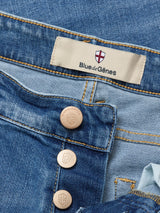 Close-up of Vinci Pala Used V2 Jeans - Denim Blue by Blue de Gênes, highlighting blue comfort stretch denim, copper shield-embossed buttons, and a fabric label with a red cross and the Blue de Gênes brand name sewn on the waistband.