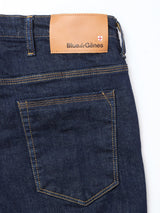 Close-up of the back of Repi Dai V Dark V2 Jeans by Blue de Gênes in dark blue denim, featuring yellow stitching and a brown leather waistband patch with the brand name and a small white and red shield logo.
