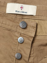 Close-up of Paulo Pavia 1 V2 Chino - Fudge by Blue de Gênes, made from cotton stretch fabric with shield-embossed metal buttons. A fabric label above the buttons displays “Blue de Gênes” and a red cross on white.