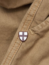 A small shield-shaped pin with a white background and red cross is attached to the seam of Blue de Gênes Paulo Pavia 1 V2 Chino - Fudge pants, adding a subtle detail to the rugged brown cotton fabric.