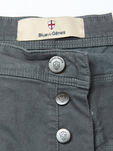 Close-up of Paulo Pavia 1 V2 Chino - Charcoal by Blue de Gênes, showing three metallic shield-logo buttons and a label with Blue de Gênes and shield emblem above the waistband. Made from cotton stretch fabric.