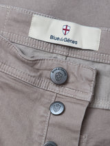 Close-up of Paulo Pavia 1 V2 Chino - Fungi by Blue de Gênes, made from light brown cotton stretch fabric. Features metal buttons and a fabric label with the Blue de Gênes name and shield logo; buttons also display the brand name and shield design.