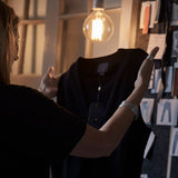 A person with long hair and bracelets holds a black sleeveless top under a glowing bulb, against a backdrop of fabric swatches and a doorway. The Merino wool knit waistcoats from Blue de Gênes feature corozo nut buttons, including the Zorba Knit Waistcoat in Navy.