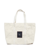The BDG Canvas Bag - Off White by Blue de Gênes is a large, beige tote perfect for everyday use. It features two sturdy handles and a front black square patch with a striking white design or text against a white background, making it a versatile accessory for any occasion.